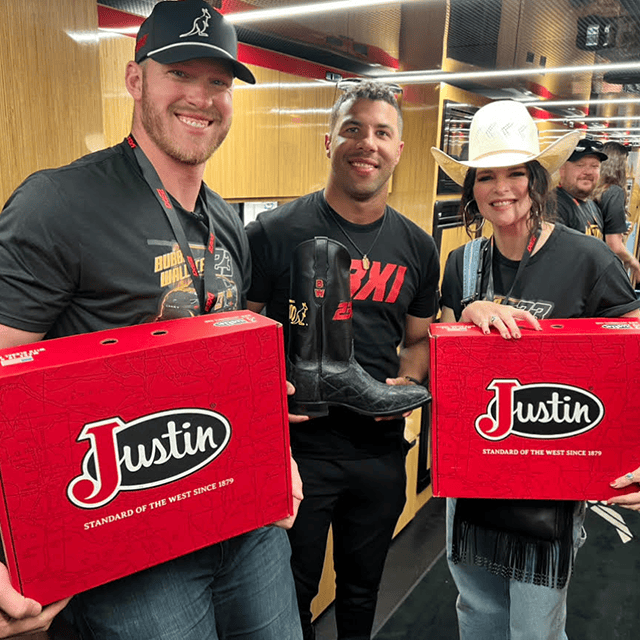 Alltroo Co-Founder Kyle Rudolph on the left holding a red Justin boot box, posing with 23XI Racing Driver Bubba Wallace, who is holding a black custom boot wearing a black t-shirt, standing next to Jenna Paulette on the right who is also holding a red Justin boot box.